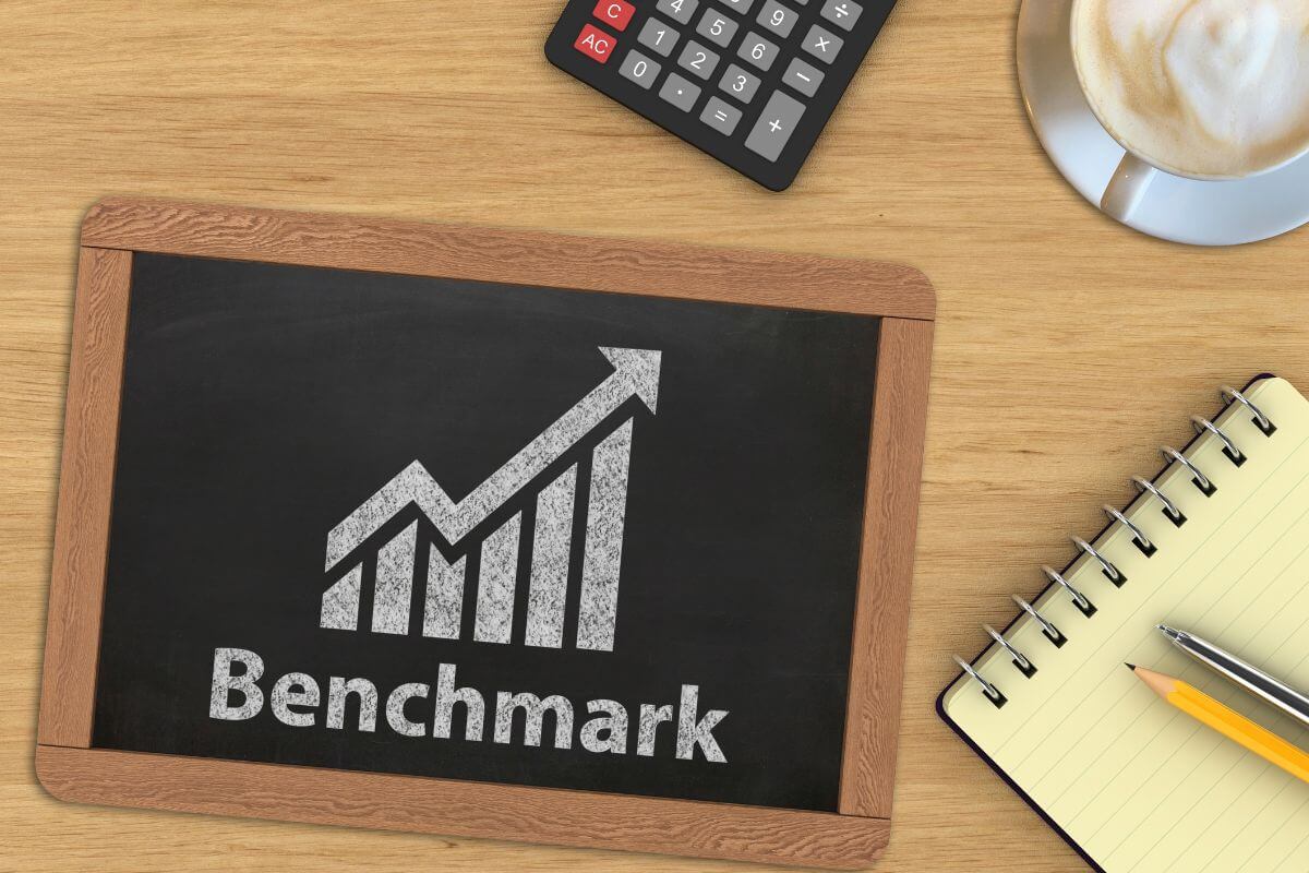 The picture tells about a calculator, a coffee, a notebook, a pen, and a blackboard with a note and graph referring to what benchmarks should be used to measure financial progress that helps you stay on track for the future.