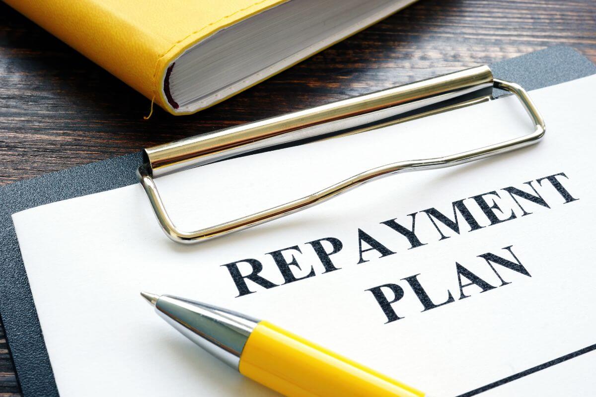 The image shows a notebook, a pen, and a clipboard with a piece of paper and notes which refer to the role of a debt repayment plan in savings strategy, repaying debt frees up money that you can redirect toward savings.
