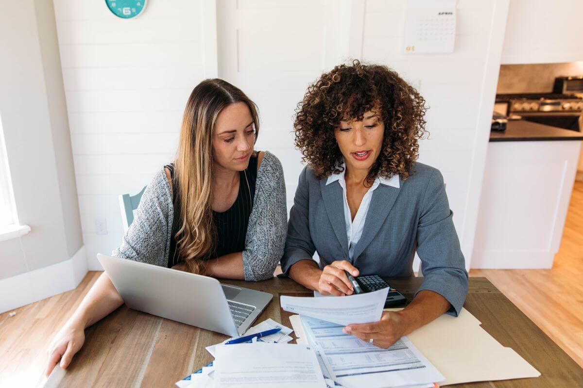 The picture shows papers on the table, a laptop, a calculator, and a financial advisor explaining the piece of paper to her client indicating that if should consider professional advice to better understand savings goals, which can help her make smarter financial decisions.