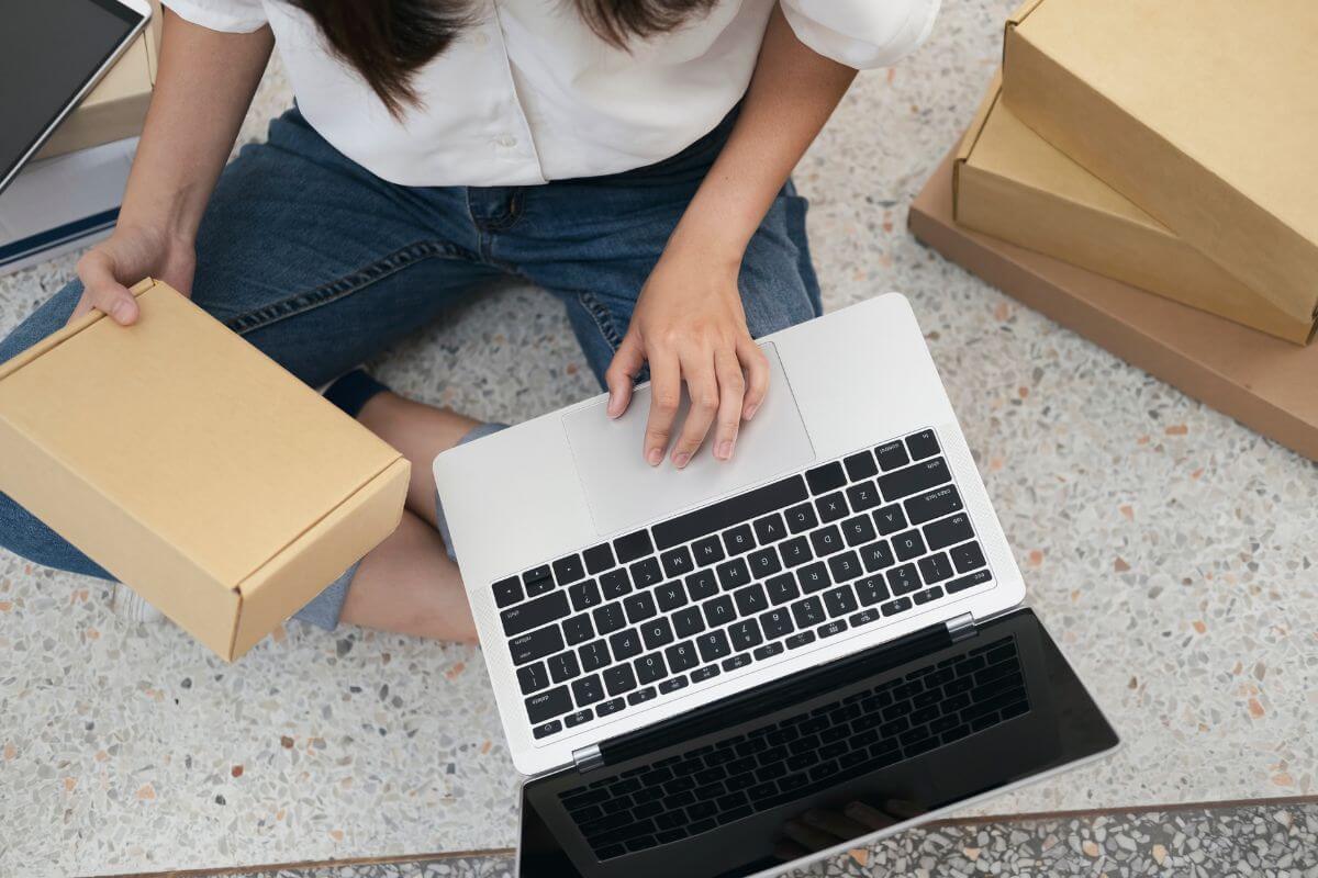 The picture tells about boxes and a girl using a laptop while holding a box which means online resellers earn a great income by flipping items on platforms that can become a consistent source of income.