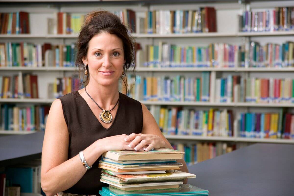The picture shows books from behind and a girl piling books on the table referring to the librarian earning good pay by managing collections and helping others find resources.