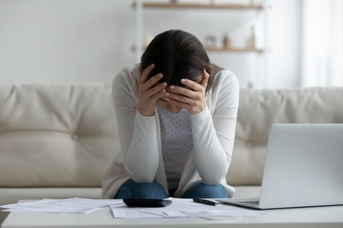 The image shows papers on the table, a laptop, a pen, a calculator, and a frustrated girl sitting on the couch which means acknowledging your mistakes can help you move forward with purpose.