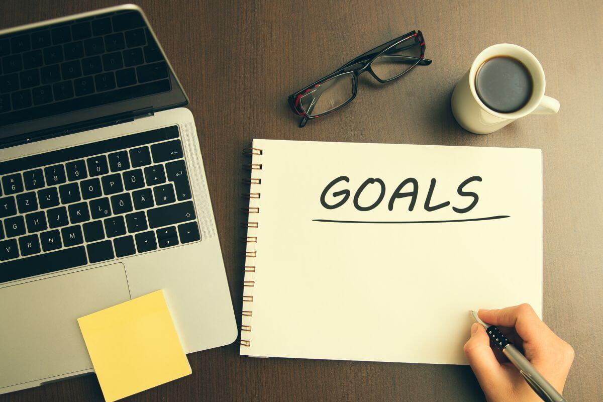 The photo of a coffee, an eyeglass, a laptop, a sticker note, and a girl's hand writing on a notebook means to set clear and achievable financial goals to keep you motivated.