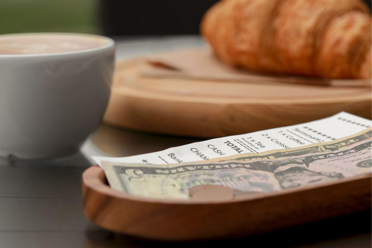 The photo tells about bread, coffee, a receipt, and dollar bills on a wooden plate referring to if you can't afford tipping, then stop eating out and consider cooking at home options instead.