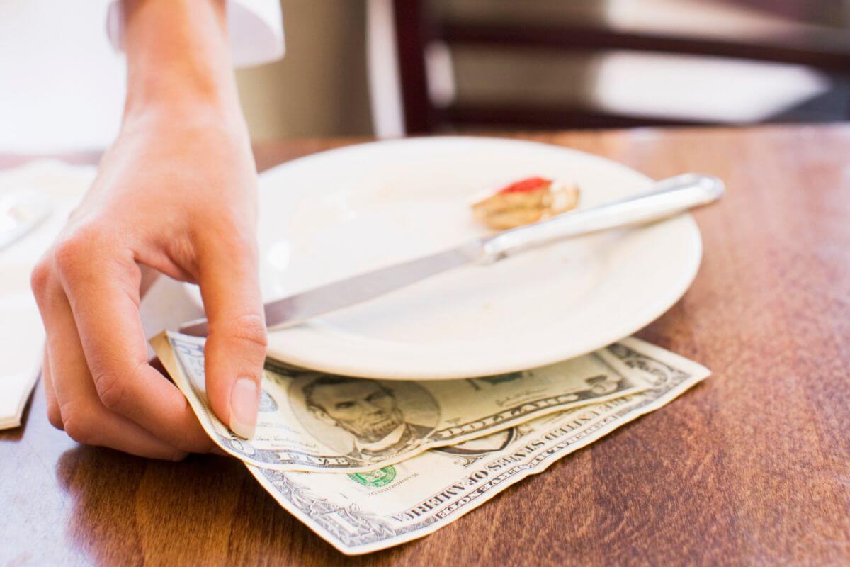 The photo tells about an empty plate with a knife and a girl's hand picking dollar bills beneath it indicating to consider tipping less if the service is horrendous and still acknowledging their work while signaling room for improvement.
