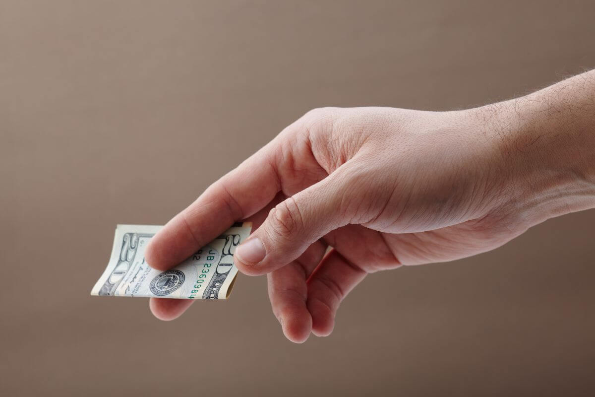 The image shows a man's hand giving a dollar bill which means digging dipper into the tipping etiquette helps avoid awkward moments and shows respect for the work being done.
