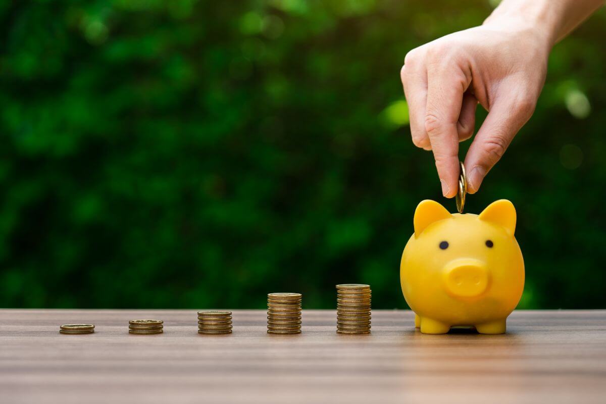 The image shows coin stocks and a man's hand dropping coins into a piggy bank indicating to start saving even a small amount that adds up over time.