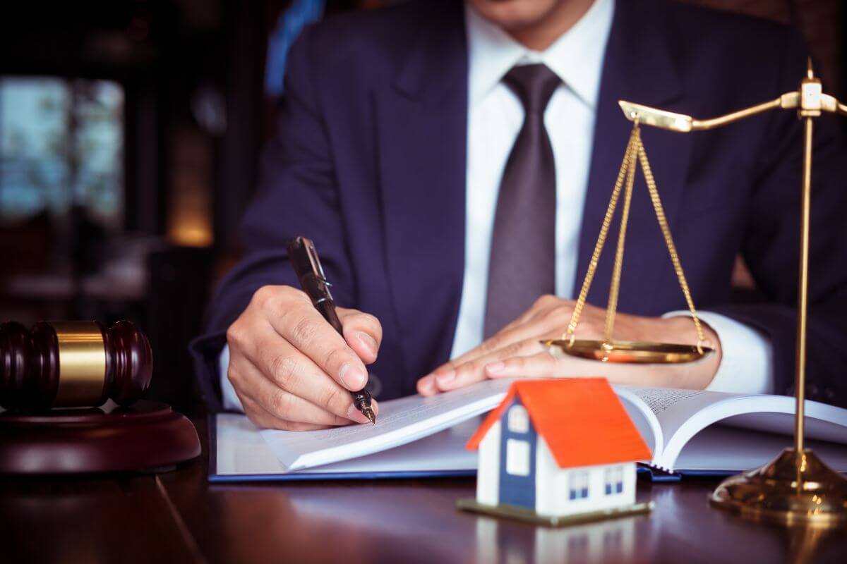 The picture tells about a gavel on the table, a mini house figure, a weighing figure, and a man's hand signing on the book which means consulting an estate planning attorney for tailored advice ensures your plan is structured to meet your specific needs.