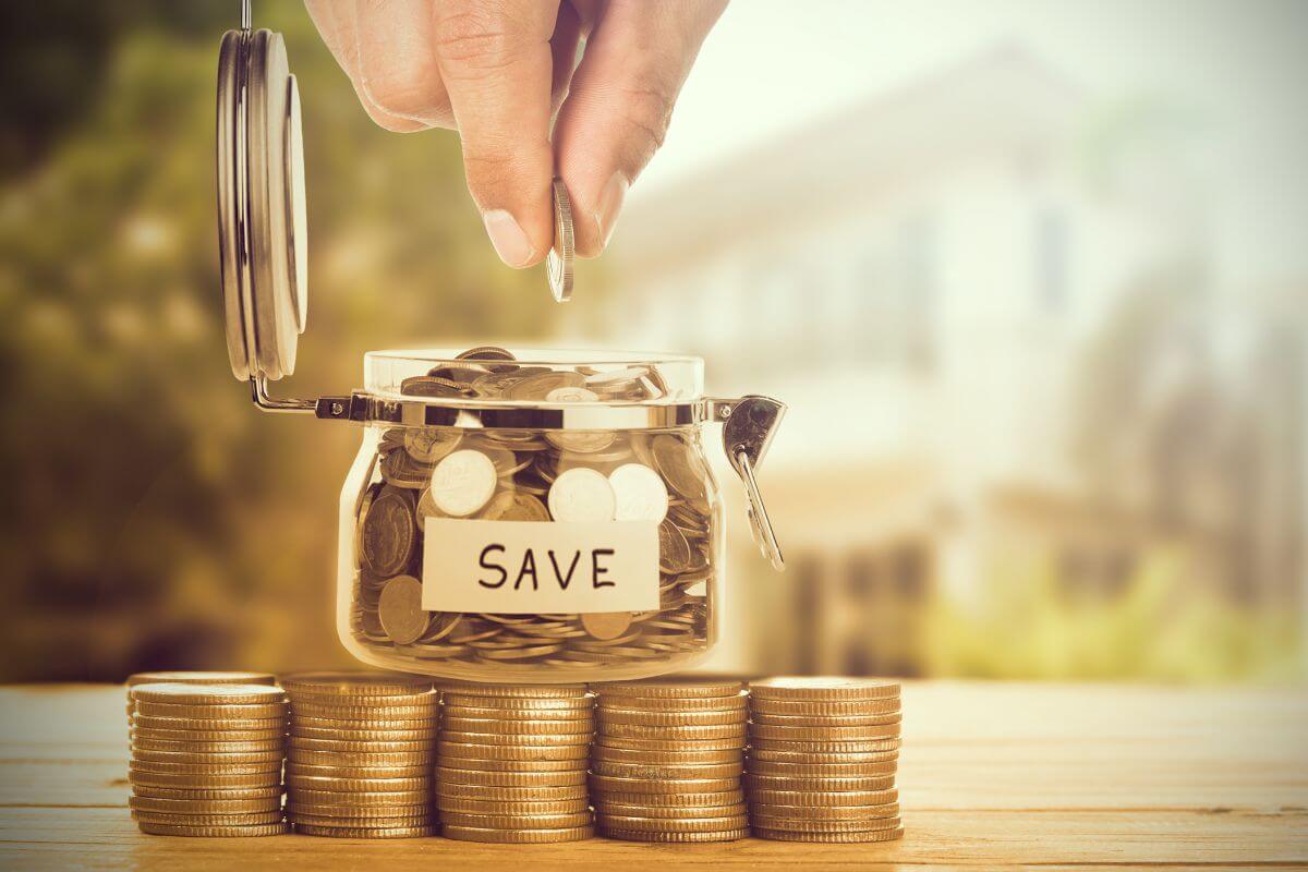 The photo tells about coin stocks, a jar filled with coins and a note, and a man's hand dropping a coin referring to automated savings and investment contributions, this removes the temptation to spend that money instead.
