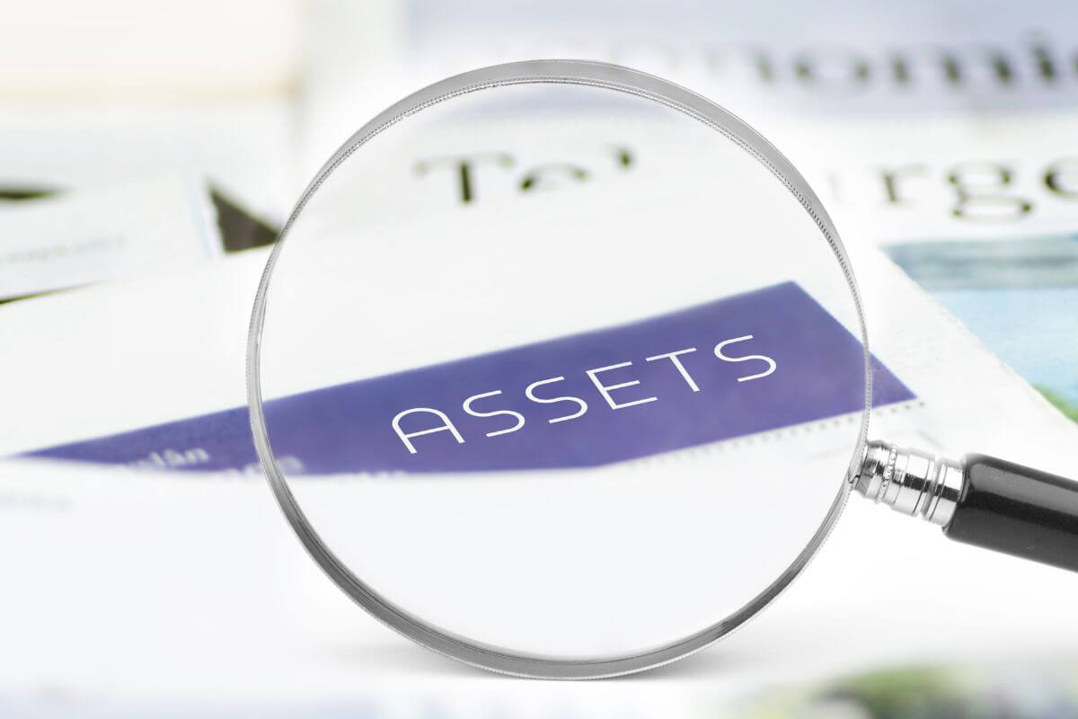 The image tells about a magnifying glass focusing on a paper with notes referring to assets which are things you own that hold financial value.