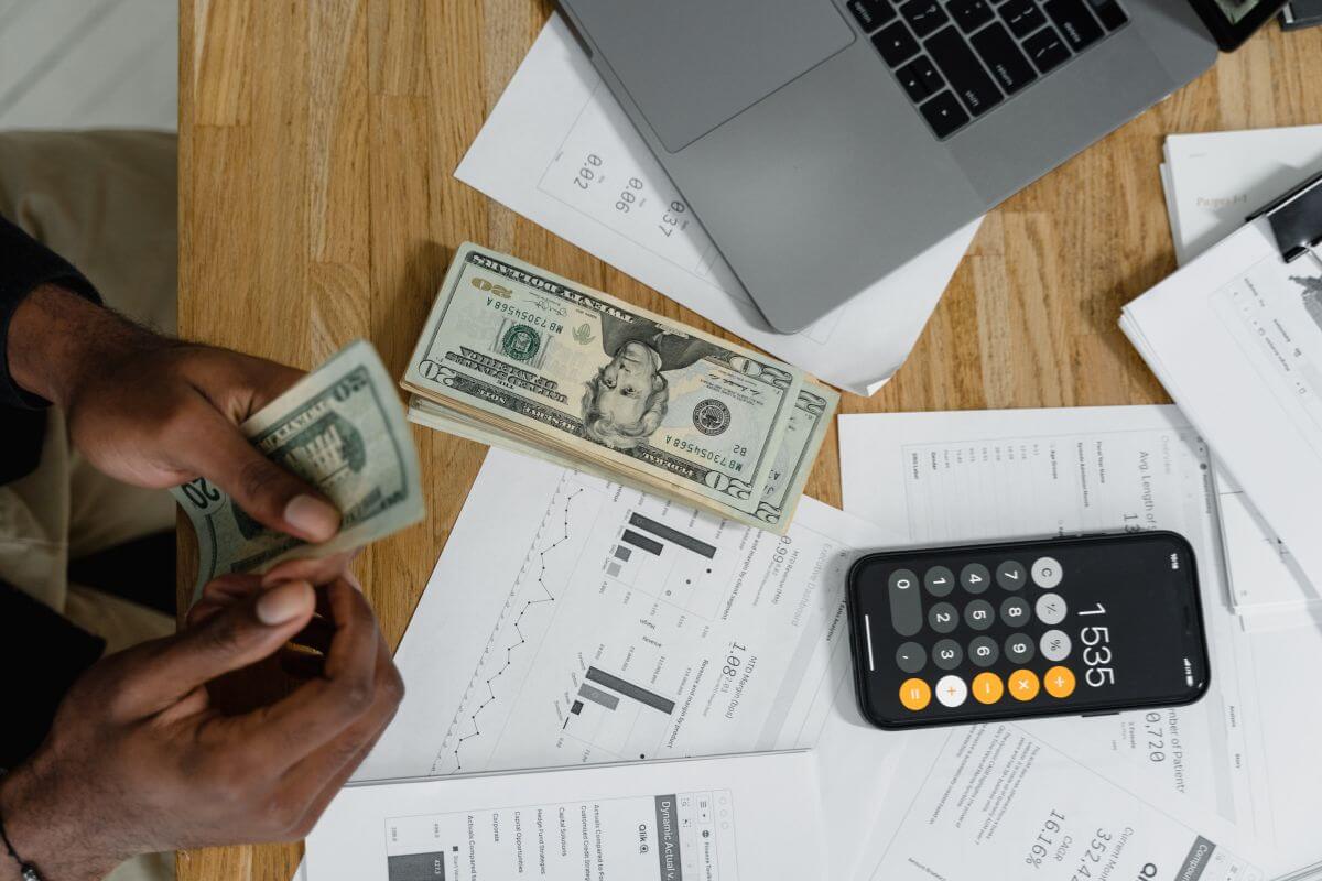 The picture shows a phone with a calculator, dollar bills on the table, papers with graphs and numbers, and a man's hand counting money which means the importance of tracking net worth shows how healthy your finances are at any moment.