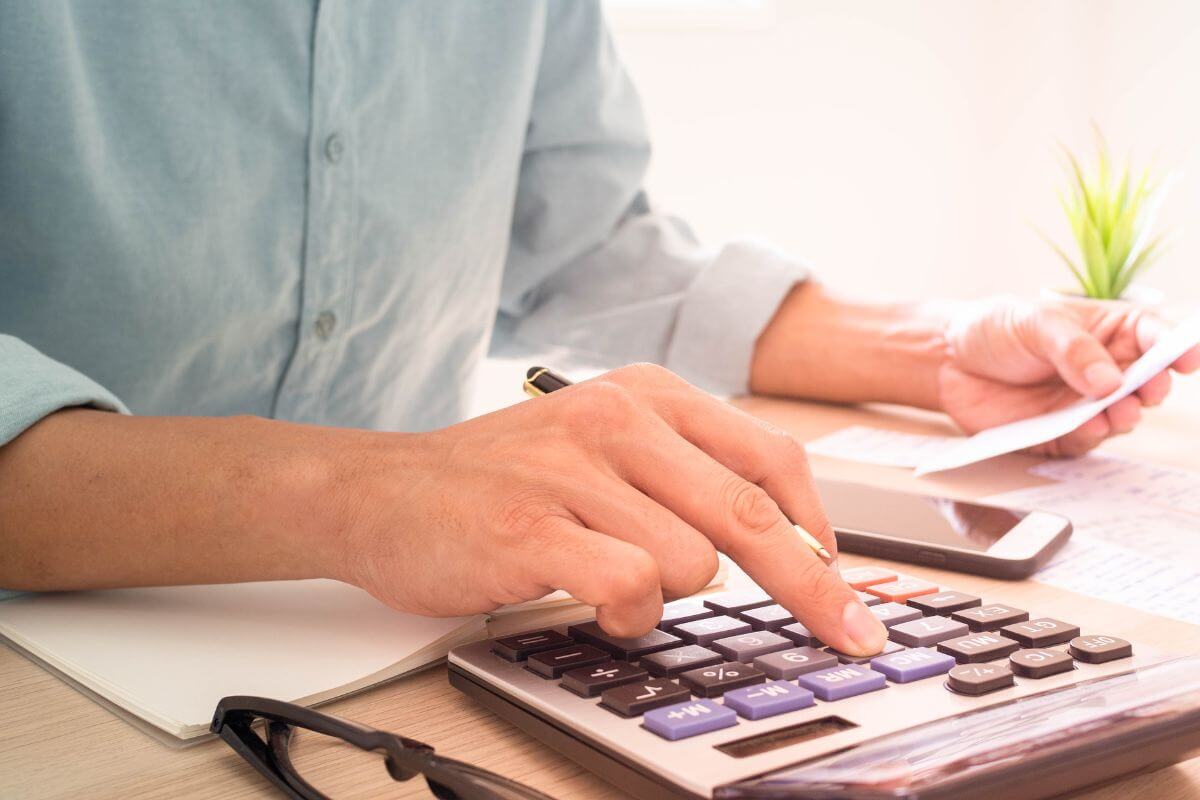 The picture tells about an eyeglass, papers on the table, a phone, and a man's hand calculating while holding a piece of paper indicating that regularly calculating and updating net worth helps you track your financial progress.