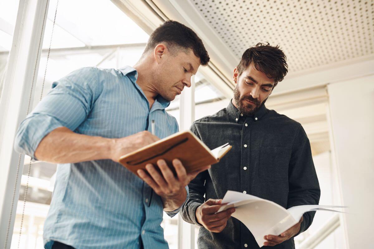 The photo tells about a man holding a notebook looking at the paper of the other man which means to avoid comparing your net worth to others can distract you from your own progress.