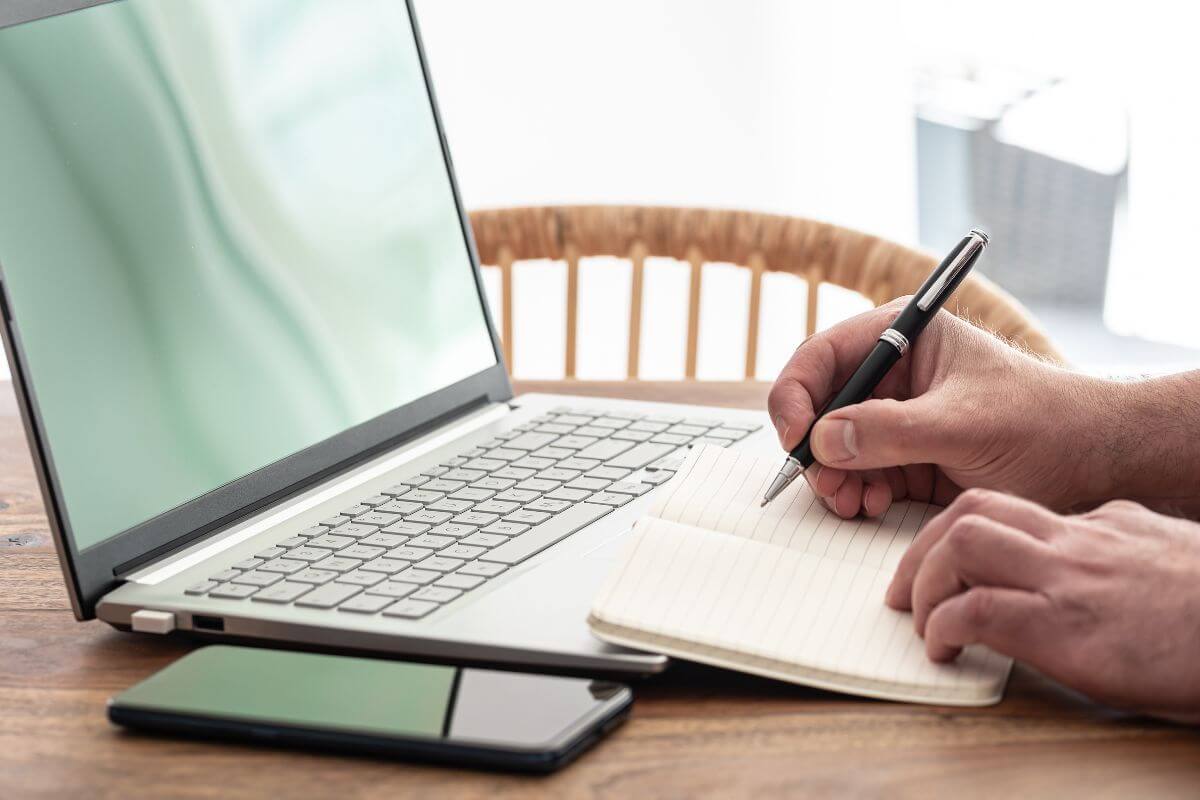 The picture tells about a phone, a laptop, and a man's hand and writing on the notebook which means to take the proper steps to build your net worth that can help you build a strong financial future.