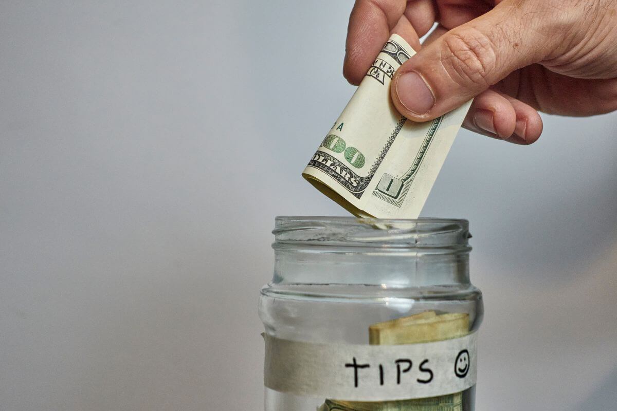 The image shows a jar filled with dollar bills and a note. And a man's hand dropping a dollar bills into it which means you should still tip to show appreciation for good services and is essential for service workers.