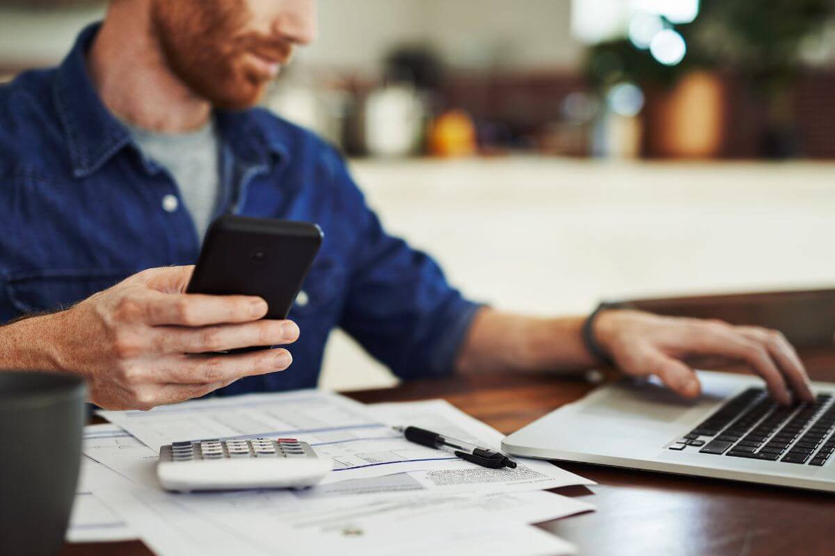 The picture of papers on the table, a calculator, a pen, and a man holding a phone while using a laptop means using tools or apps to track your progress to see how close you are to your goals and help you reach your nest egg target faster.