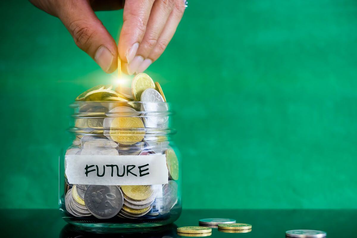 The photo of coins on the table, a jar filled with coins and with a note and a man's hand dropping a golden coin into it refers to start transforming your financial future which makes a big difference in building a more secure financial future.