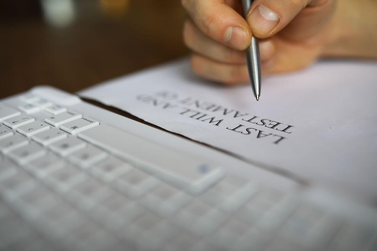 The picture shows a keyboard, a piece of paper with notes, and a man's hand holding a pen, which refers to including a backup executor to ensure there is someone ready to take charge and follow your wishes.