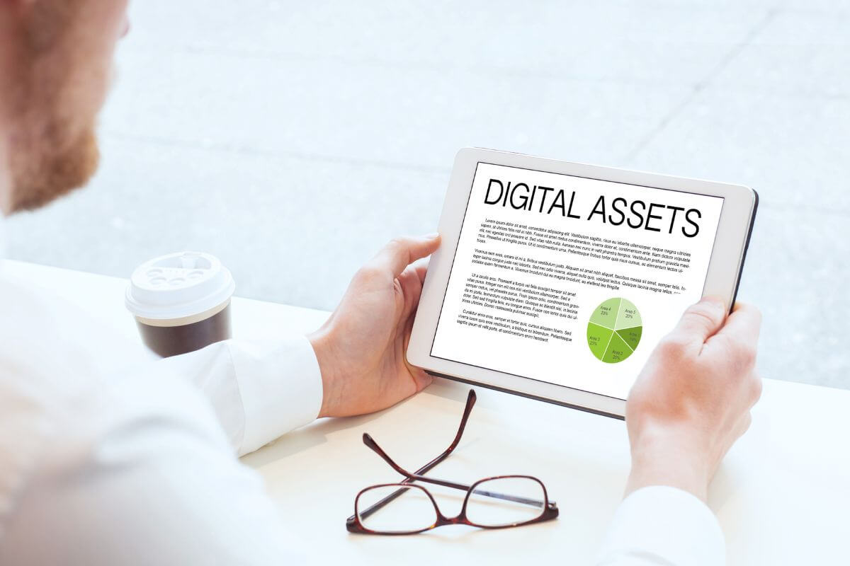 The picture shows coffee, an eyeglass, and a man holding a tablet with a screen display indicating that specifying digital assets helps your loved ones handle things better.