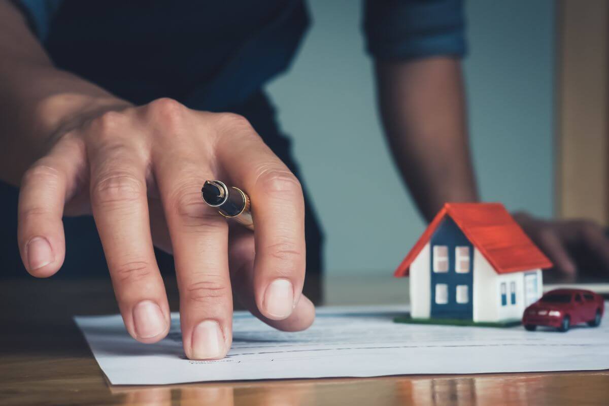 The photo shows a mini house and car, a piece of paper on the table, and a man's hand holding a pen which means providing details for real estate helps ensure they're passed on according to your wishes.