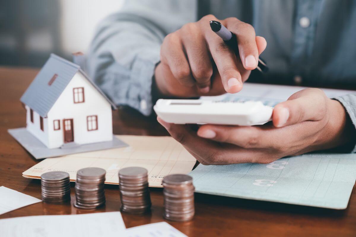 The picture shows coin stocks, bank books on the table, a mini house, and a man's hand calculating which refers to downsize housing can free up funds for savings or everyday costs.