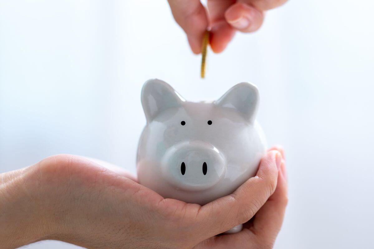 The photo tells about a girl's hand holding a piggy bank while dropping a coin into it which means using a mini-saving challenge to help create a routine of saving and show how little actions can add up.