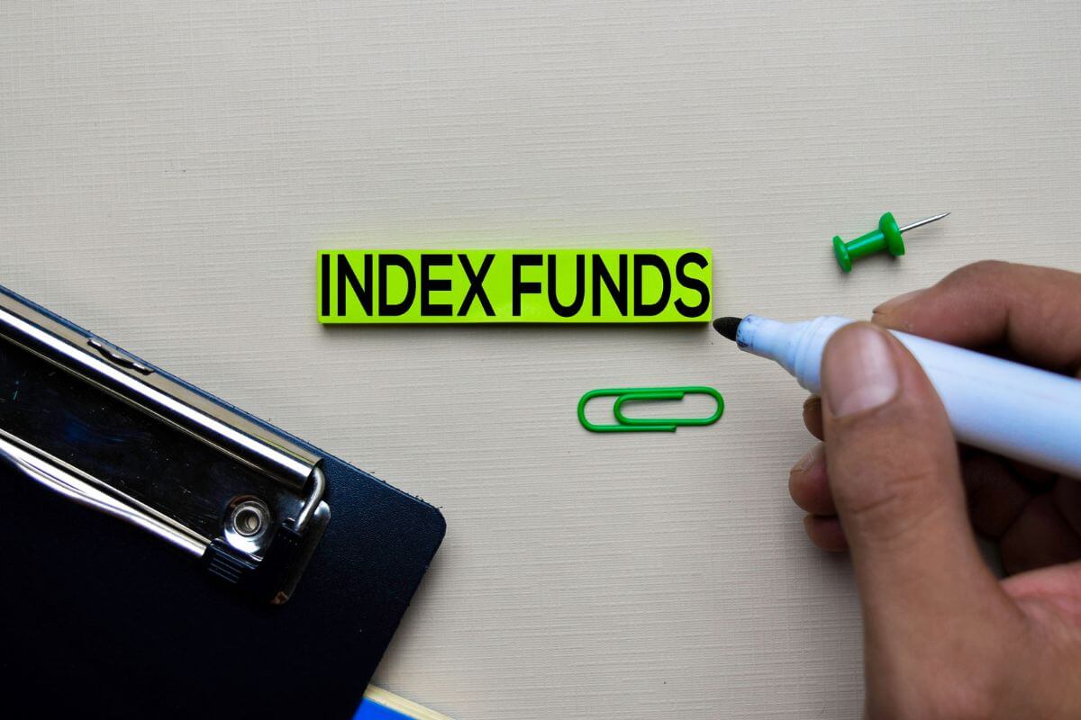 The photo tells about a clipboard, a pin, a man's hand with a marker, and a piece of paper with notes indicating that simple investment options like index funds let you grow your money steadily while you learn about investing.