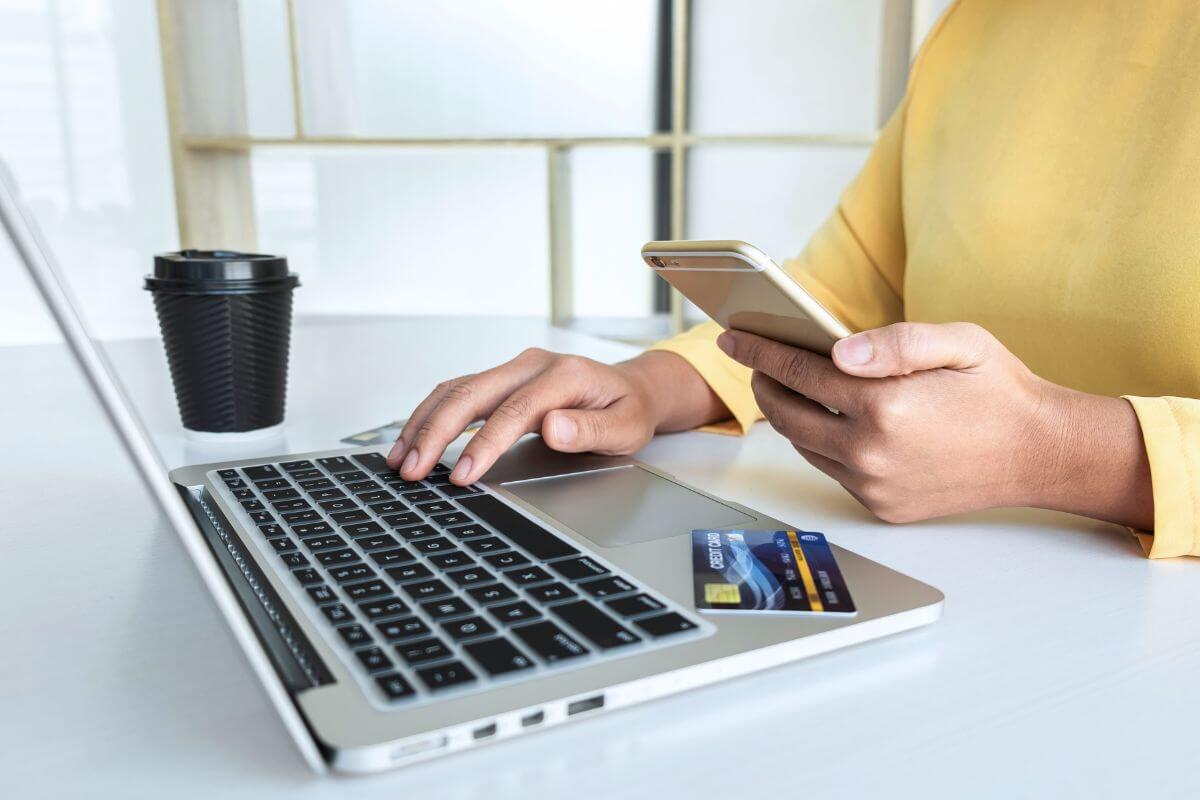 The photo tells about a coffee on the table, a card, and a girl's hand using a phone while typing on the laptop which means to open a secured credit card with a low deposit helps you qualify for better options.