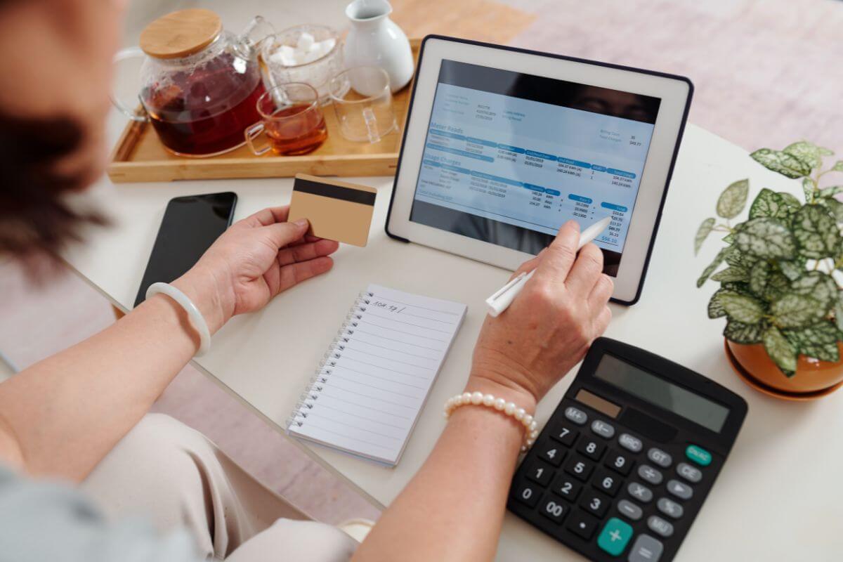 The image shows a tea pot, a plant, a calculator, a notebook, a phone, and a girl's hand holding a card while using a tablet device which means paying all bills on time helps improve your credit score on time.
