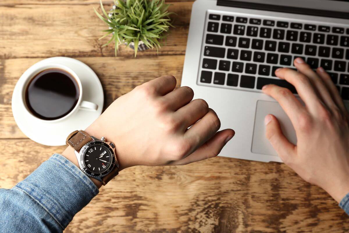 The picture shows a coffee, a plant, and a man's hand typing on the keyboard while looking at his watch referring to work in a 52-minute burst which is a great way to make steady progress without feeling overwhelmed.