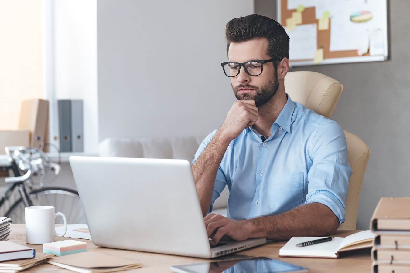 The photo tells about notebooks, a mug, a sticker note, a tablet device, a pen, and a man working on his laptop which means using temptation bundling to pair an enjoyable activity with something fun makes hard work easier to handle.