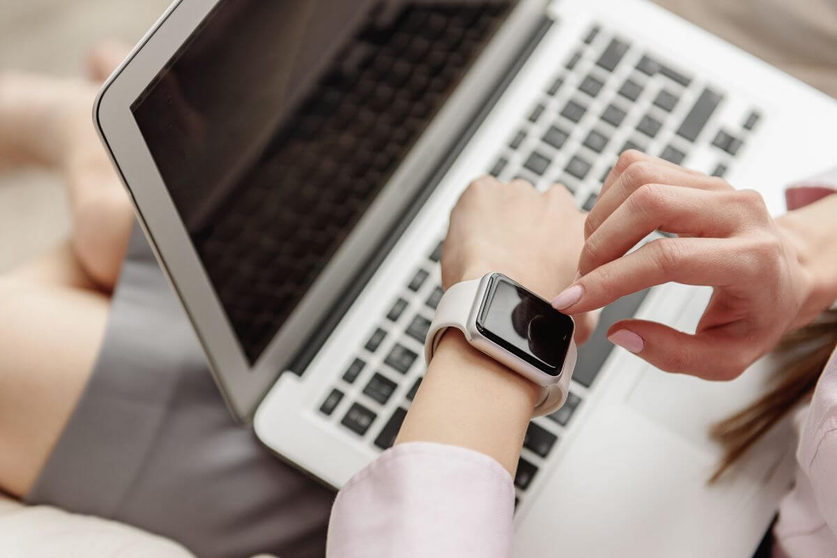 The image shows a girl tapping her watch with a laptop on her lap indicating that using your time wisely and finding productive things to do when bored helps you accomplish more even during slow moments.