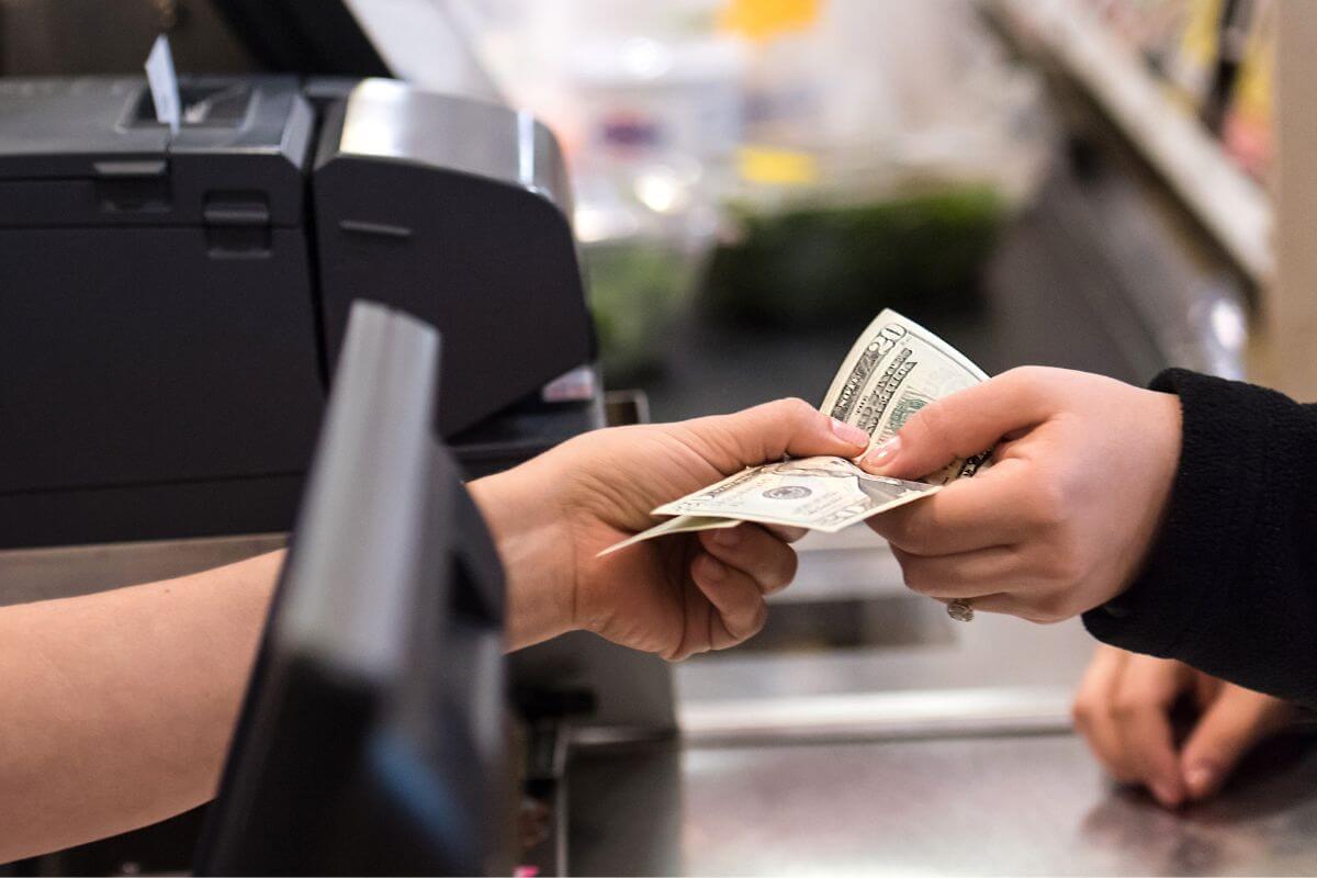 The image tells about a customer's hand giving dollar bills to the cashier's hand referring to switching to a cash-only budget to keep you mindful of your finances and ensuring that every dollar goes toward essential needs.