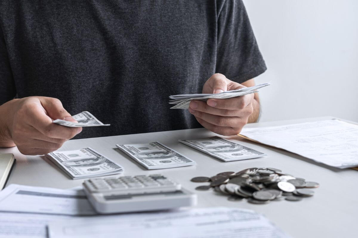 The image shows papers, coins, a calculator, and a man grouping the dollar bills referring to tracking every dollar you spend for a month can help you prioritize needs and limit splurges.