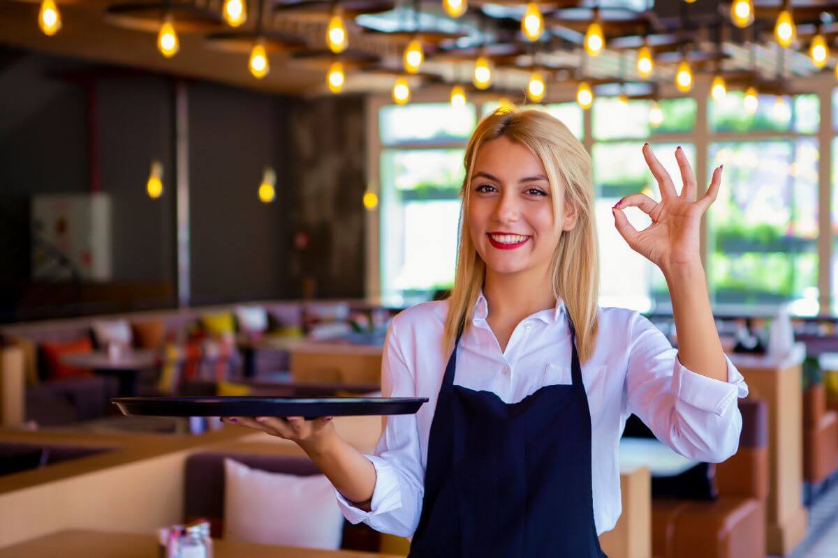 The photo shows a waitress holding a tray and doing an ok sign referring to taking on temporary gig work can help you earn extra cash fast.