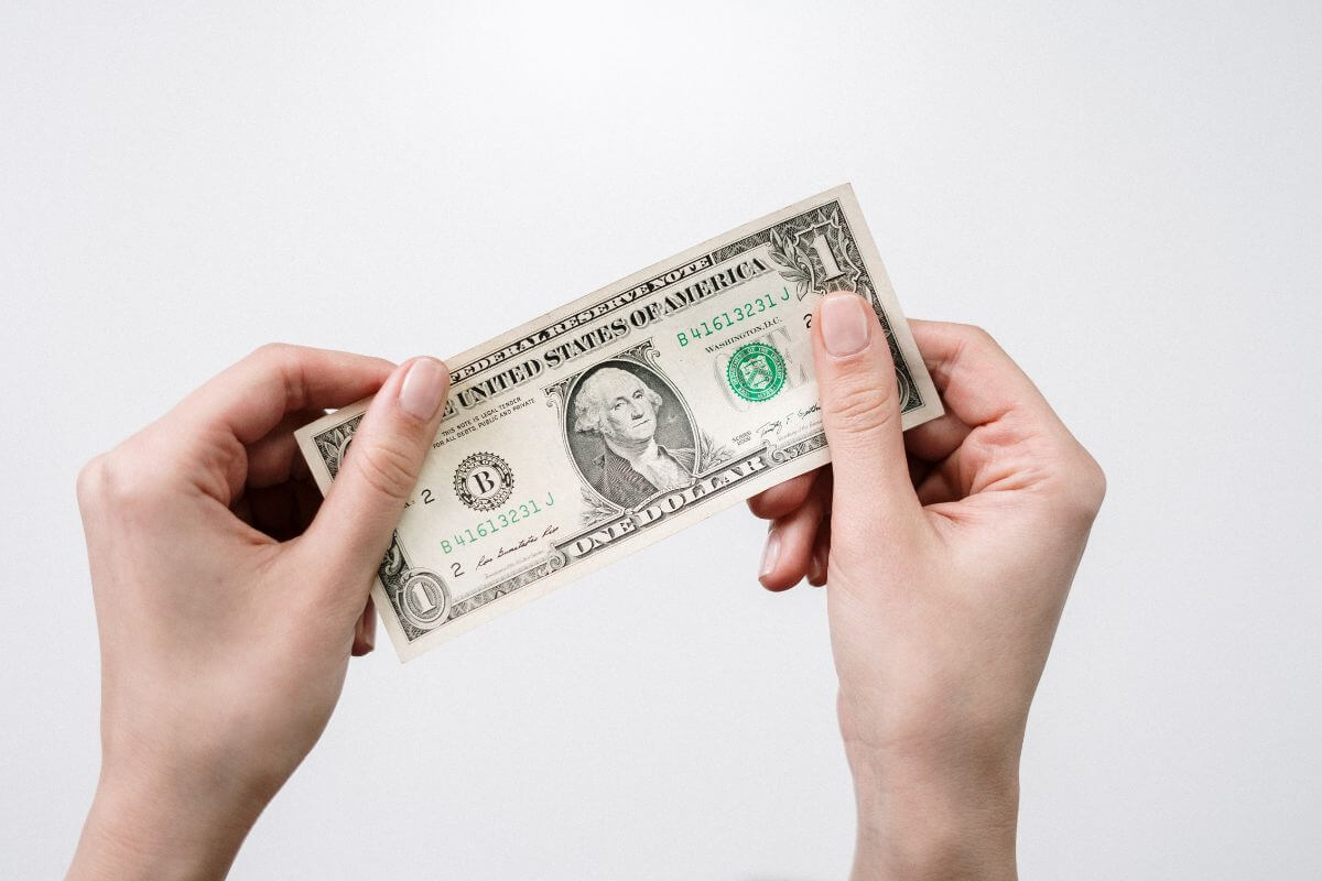 The image shows a girl's hand holding a dollar bill indicating to challenge yourself to save just $1 per day that can increase gradually the amount and save even more.