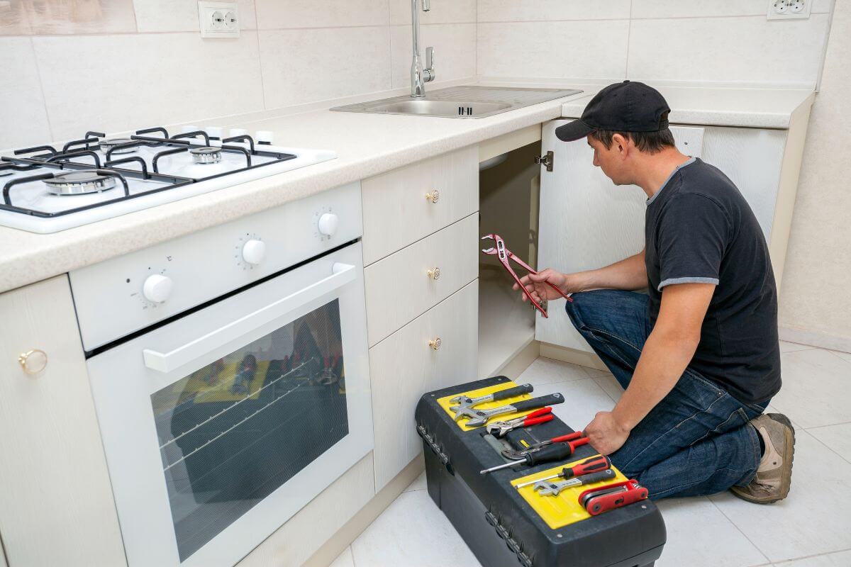 The image shows a toolbox and a man's hand repairing the plumber of the kitchen sink which means to consider bartering services for a place to stay and keep a roof over your head without spending money.