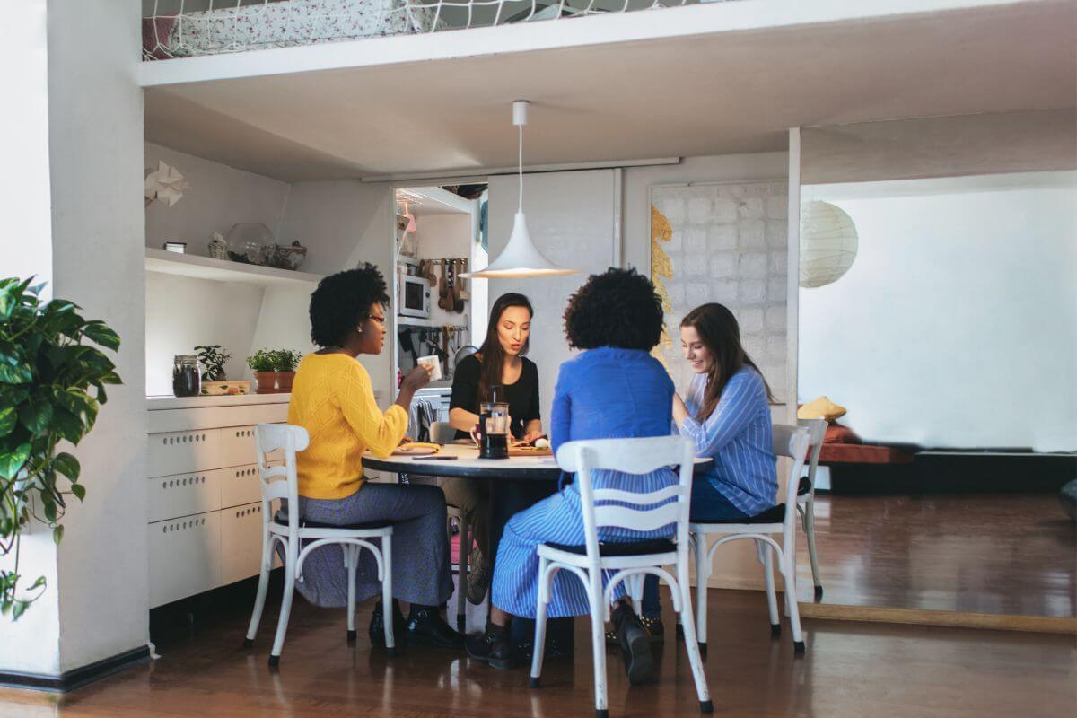 The image tells about a group of people eating at the table referring to looking into co-housing arrangements to lower your rent and meet housing needs. 