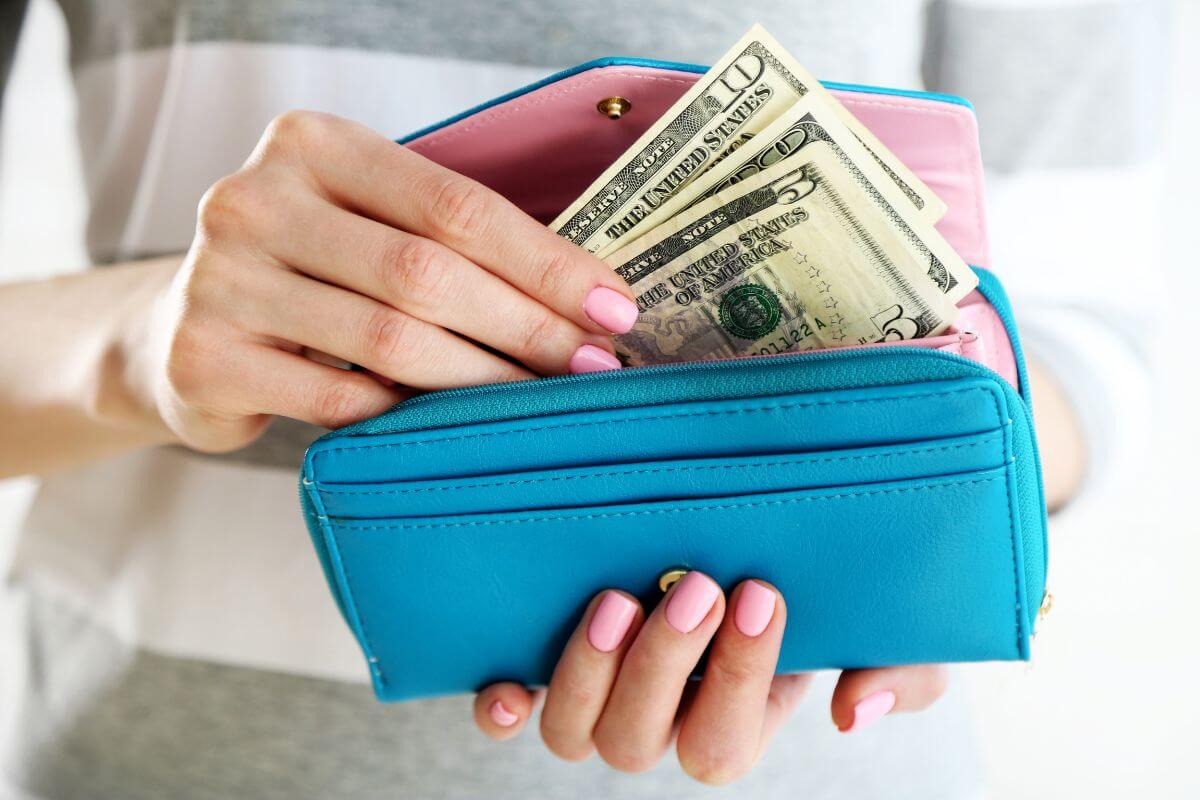 The image shows a girl's hand putting back the money inside her wallet referring to starting a no-spend challenge and seeing how much you can save by simply saying no to excess spending.