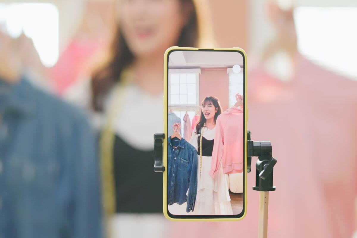 The image shows a girl holding clothes while filming herself on her phone, indicating that she can buy and sell used clothes online and turn unused items into extra cash.