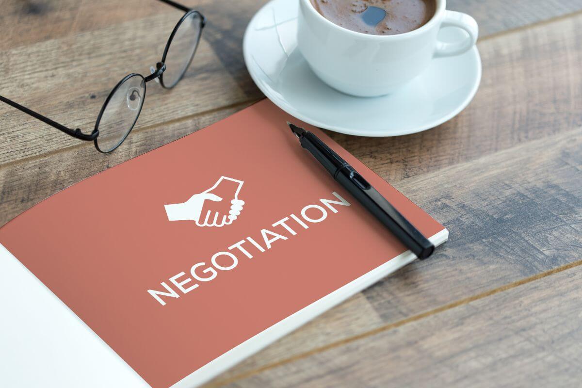 The photo shows an eyeglass, a pen, a coffee, and a book with a title and graphic shaking hands which means to learn to negotiate prices on things to get the best deals and save money.