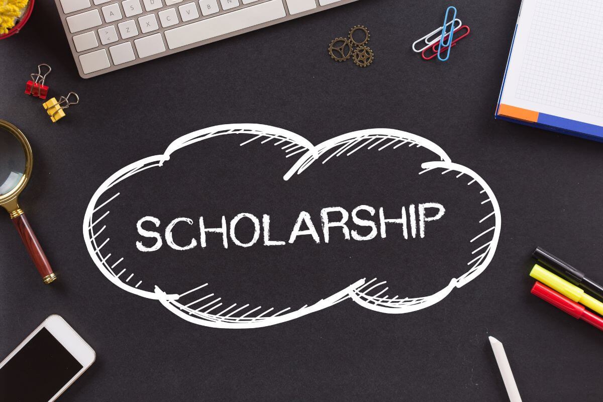 The image shows a keyboard, clips, a phone, a notebook, and a note written in chalk referring to research scholarship early to make higher education more affordable while teaching persistence and planning.