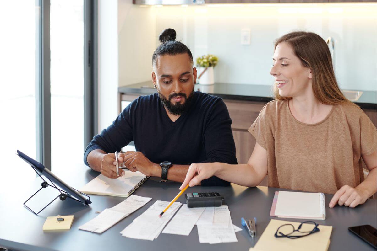 The photo tells about papers on the table, a tablet, a calculator, notebooks, a phone, and a couple pointing the paper which means to find an accountability partner can encourage each other to stick to budgets and avoid splurges.