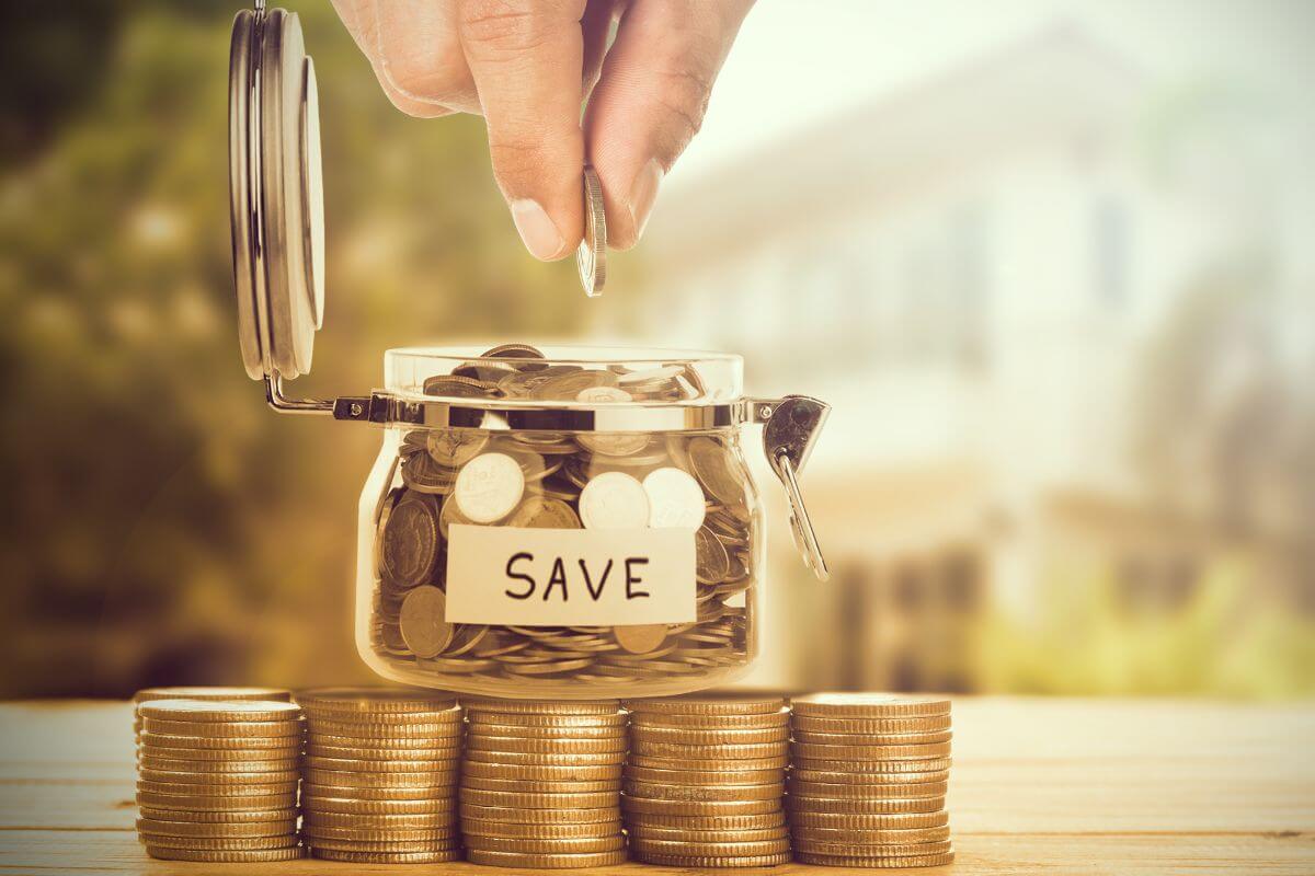 The image tells about stock coins, a jar filled with coins and notes, and a man's hand dropping a coin to it referring to making saving a game by competing with friends that can add some fun to the process while keeping everyone accountable.