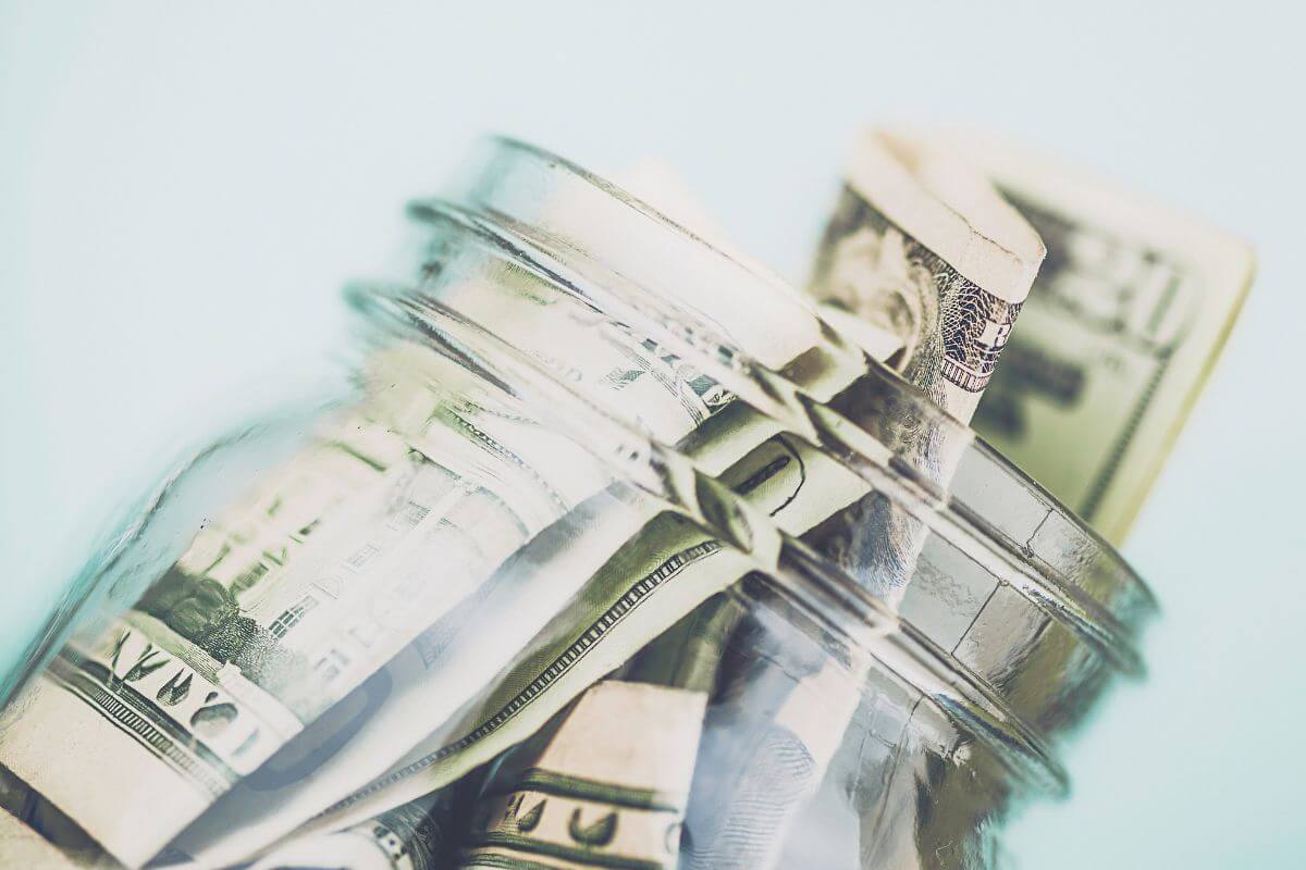 The photo of a jar filled with dollar bills means making saving visual to stay inspired and stick to your money savings plans.