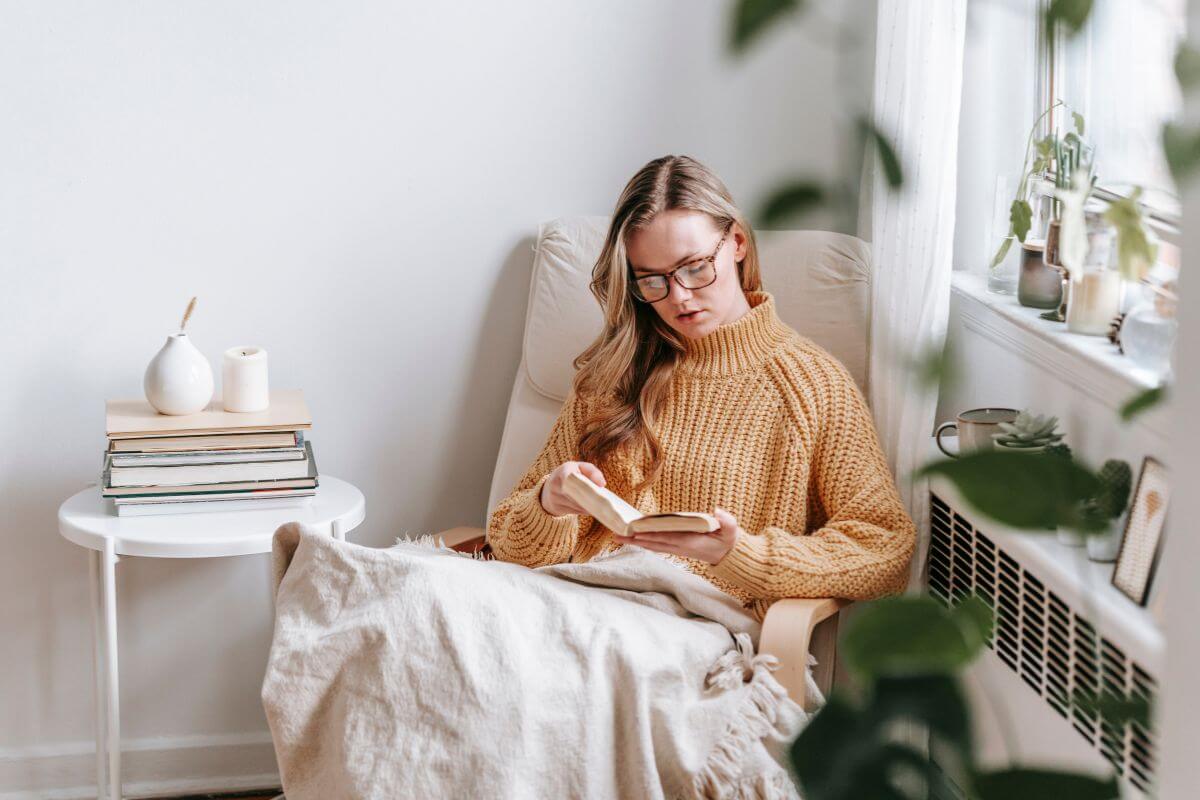 The image shows books on the table, and a girl sitting on the chair while reading a book which means a luxury house-sitter for wealthy clients is a low-pressure job that often comes with perks like staying in beautiful homes.