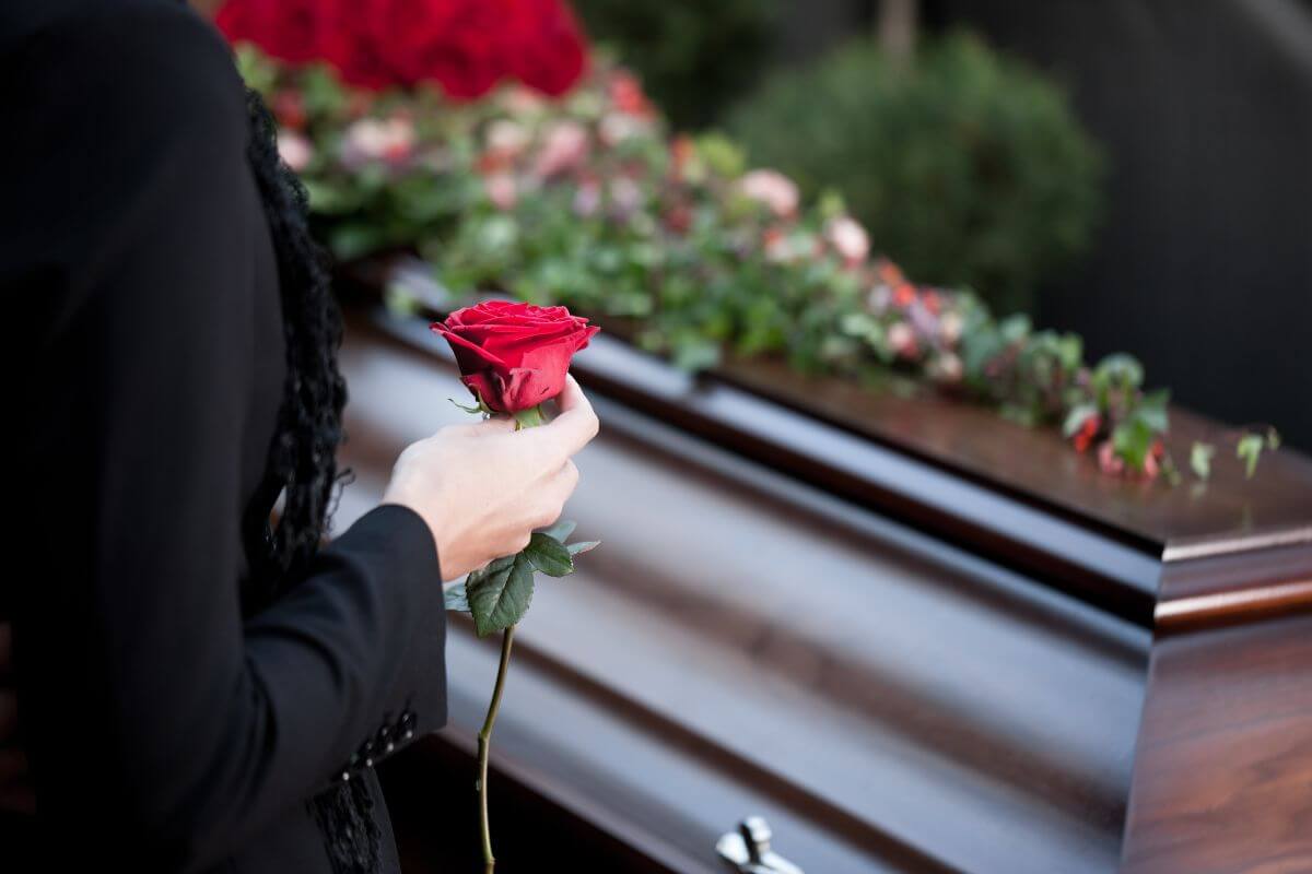 The photo shows a coffin with flowers on top and a girl in a black suit holding a rose referring to a professional mourner attending funerals that offers a way to contribute meaningfully while earning a good income.