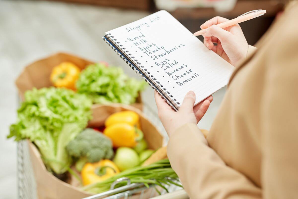 The image shows a cart with paper bags with vegetables and a girl checking her notes with a pen referring to only shops with a pre-written list keeps you from overspending and falling for unnecessary items. 