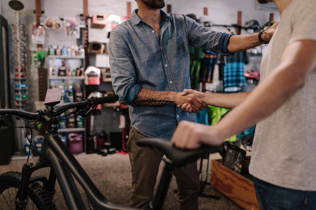 The image shows a man's hand with tattoos shaking the hand of a boy holding a bike which means to sell your bike everywhere to cut down on transportation costs and make your budget more manageable.