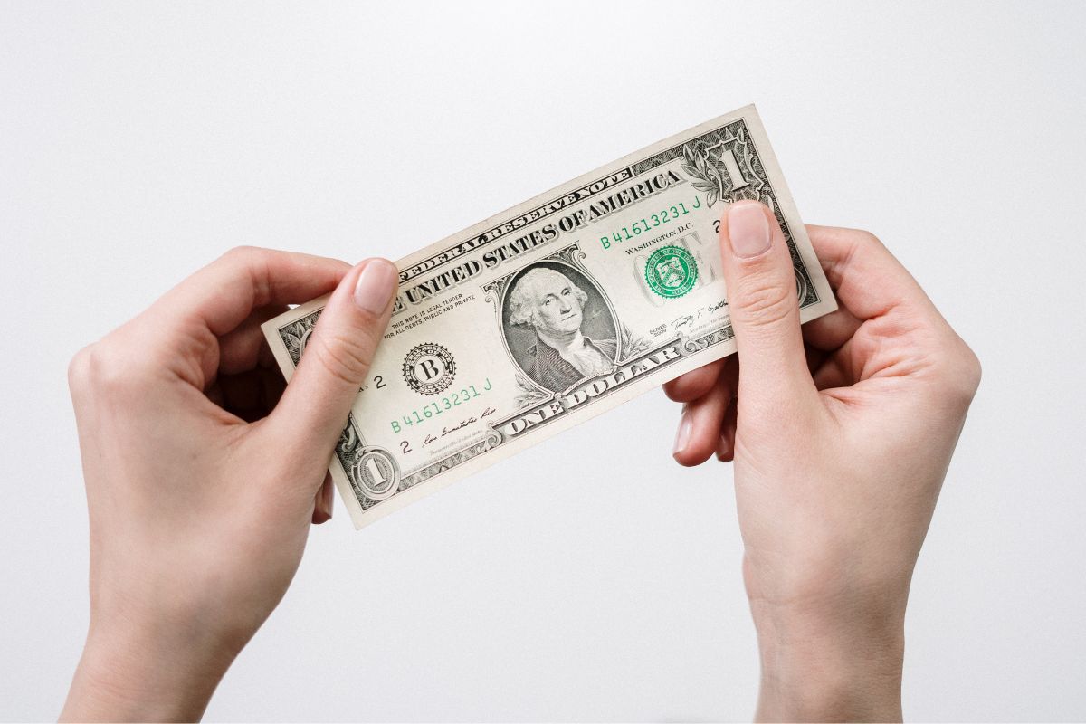 The image shows a woman's hand holding a one-dollar bill.